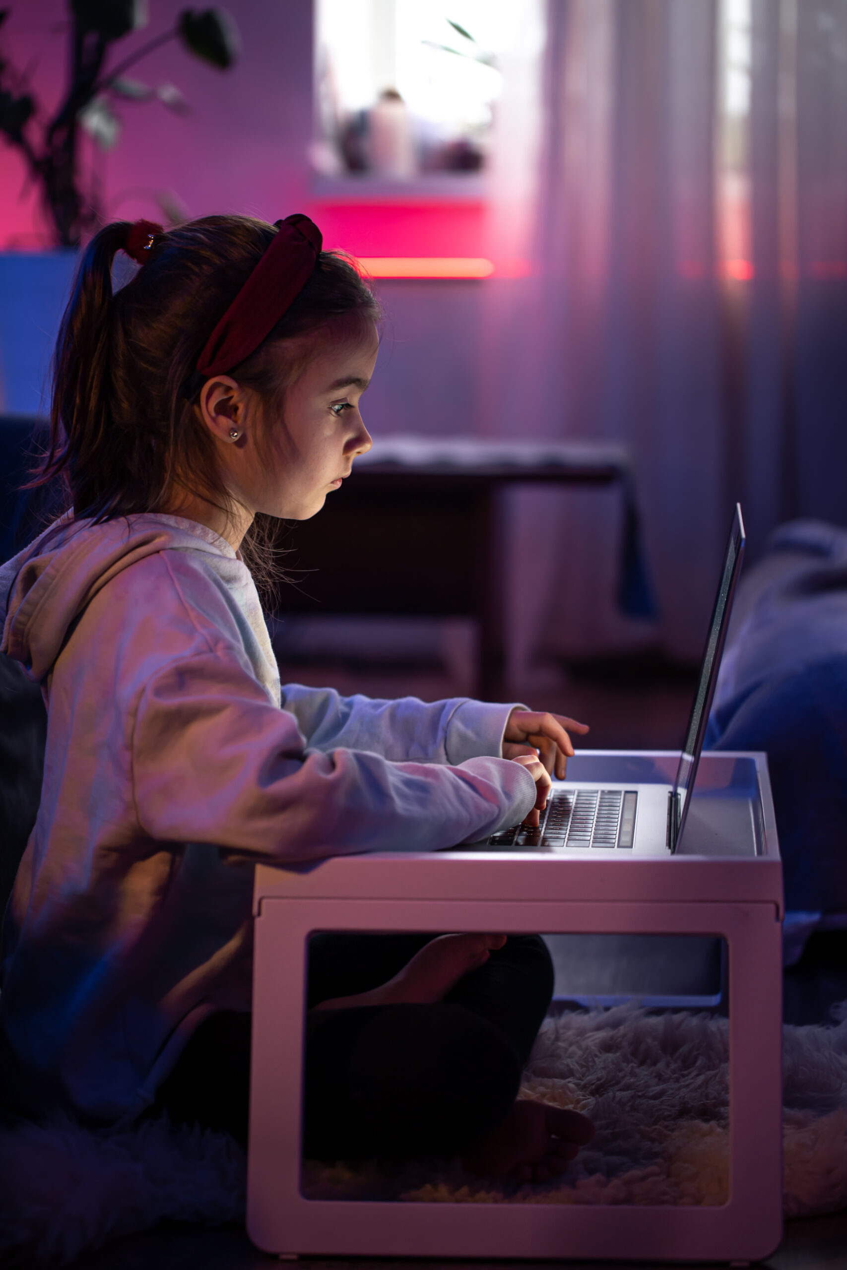 A little girl uses a laptop while sitting in a room with neon lighting.